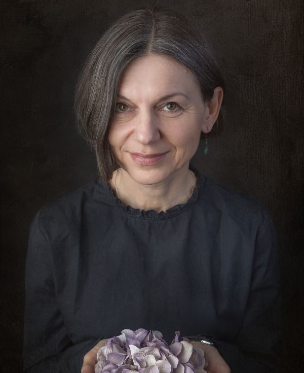 Portret kobiety en face, z krótkimi ciemnymi włosami, lekko uśmiechniętej, trzymającej kwiaty.