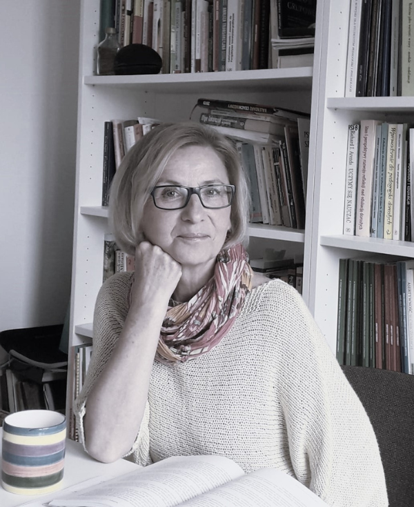 Kobieta o jasnych włosach, w okularach siedząca przy stole. Opiera policzek na prawej ręce.