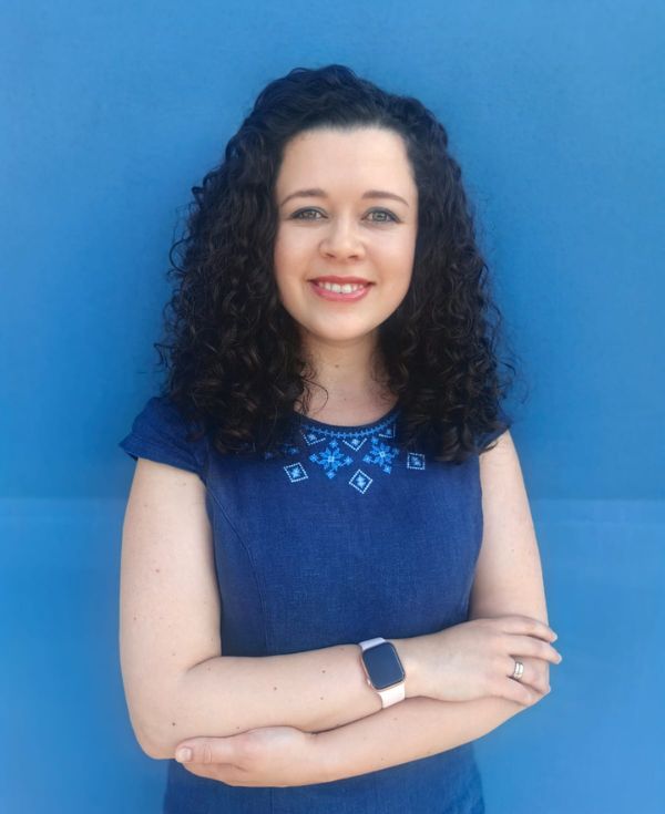 Zdjęcie przedstawia portret en face uśmiechniętej kobiety o czarnych włosach z założonymi rękami.