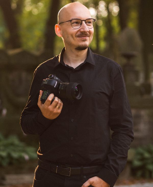 Mężczyzna w okularach ubrany na czarno z aparatem w dłoni.