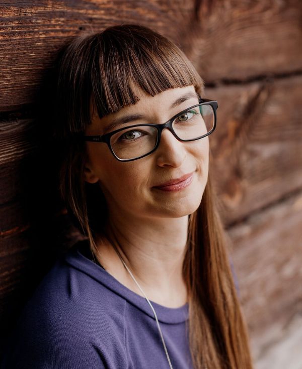 Kobieta o długich włosach z grzywką, w okularach i fioletowej bluzce